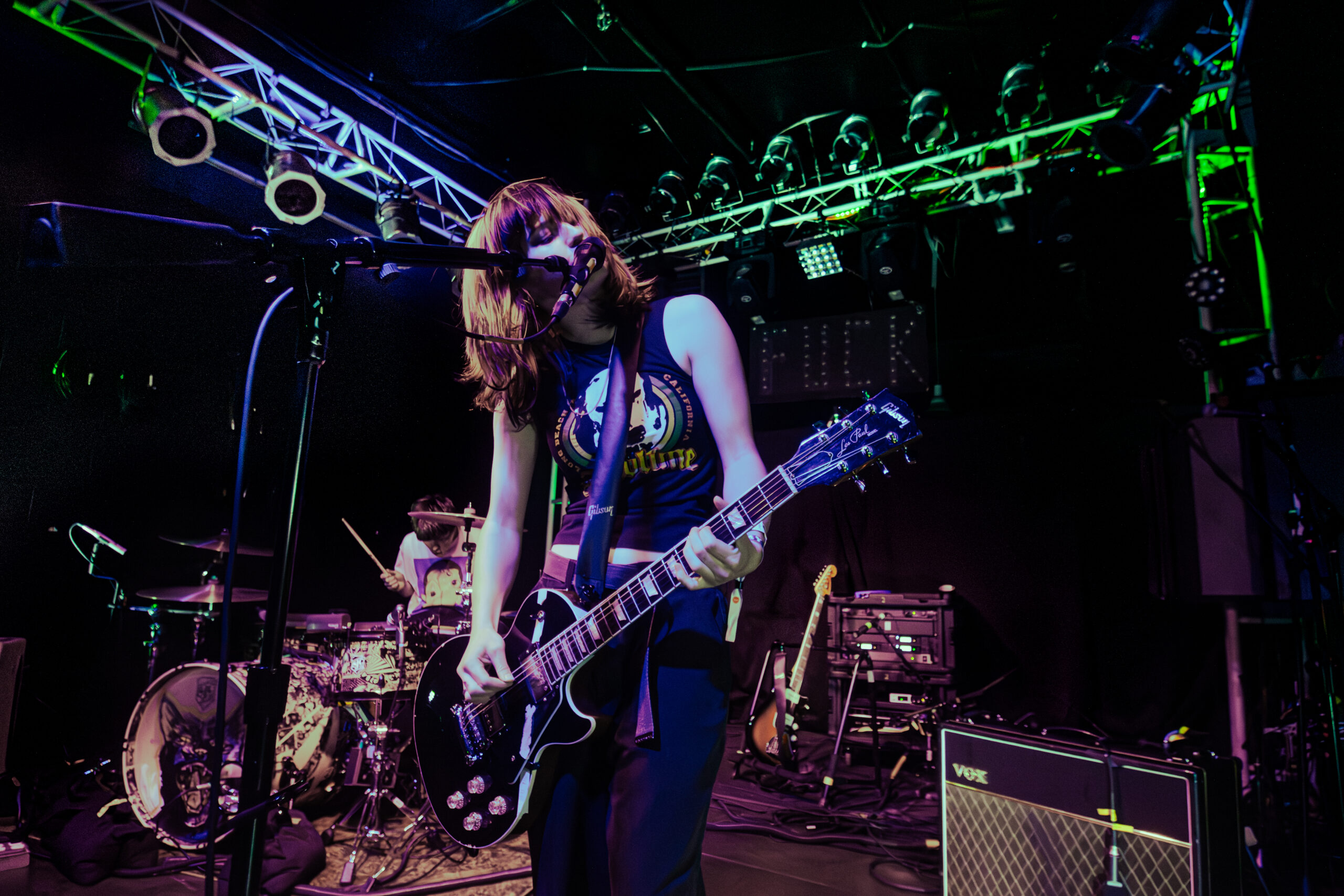 Lead singer and guitarist of RAUE performing onstage with drummer in the background.