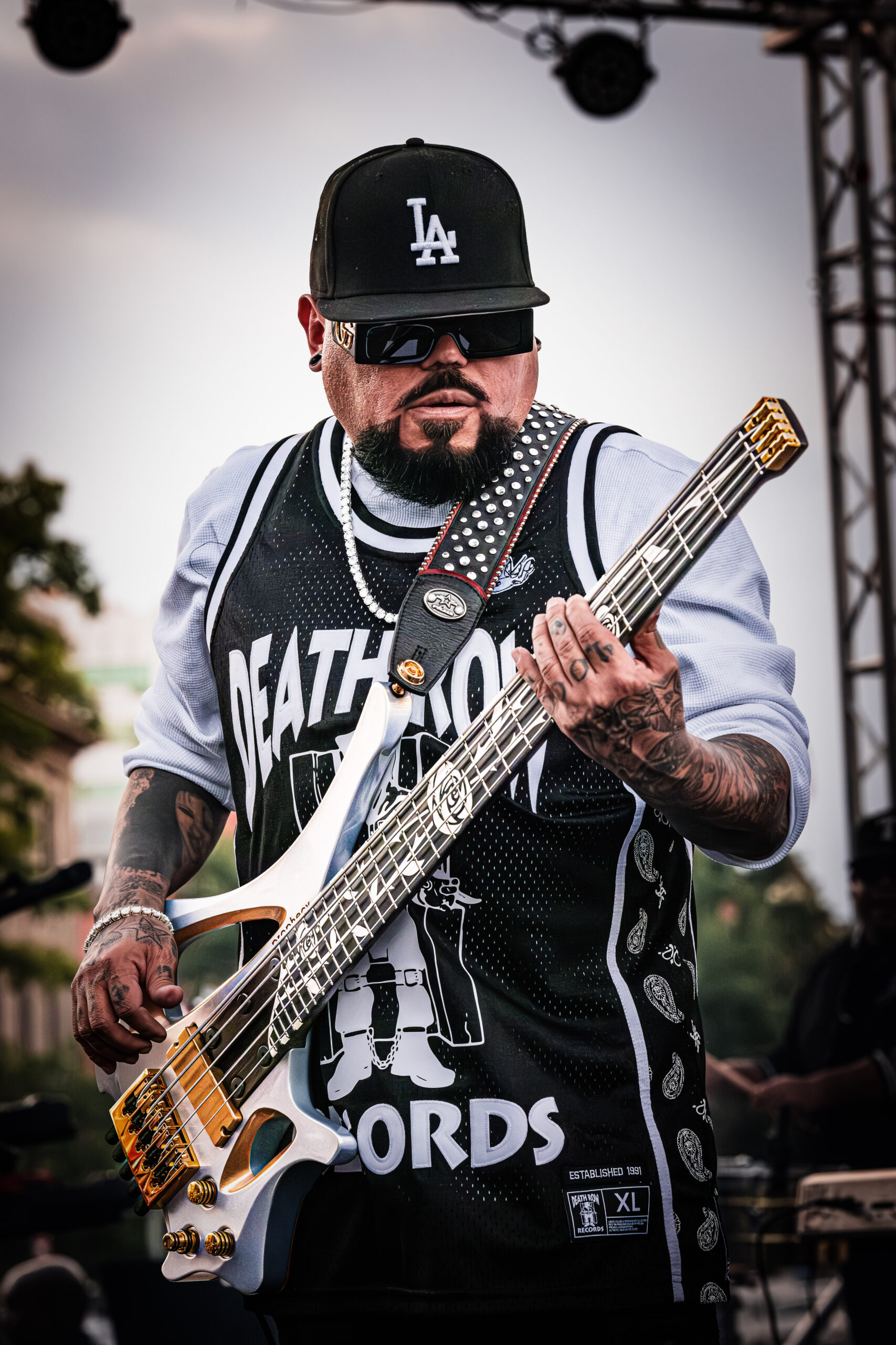 A.B. Quintanilla of the Kumbia All Starz playing the bass guitar on stage, wearing a black LA cap, sunglasses, and a Death Row Records jersey.