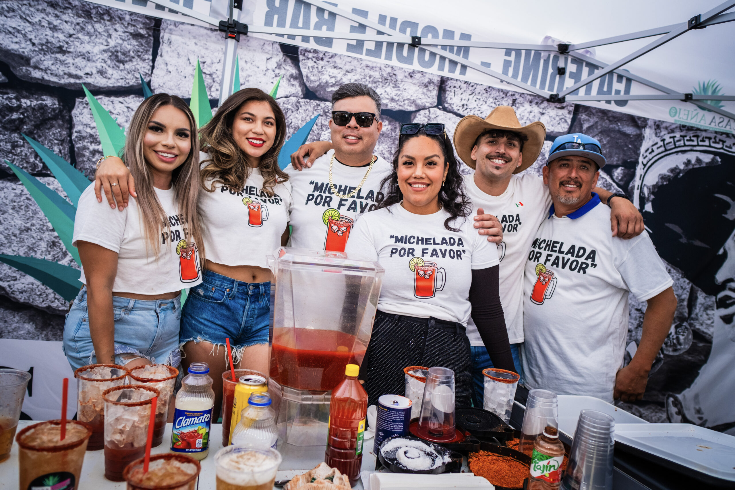 A group of six smiling people standing behind a table filled with Michelada ingredients at Latino Fest 2024, all wearing matching "Michelada Por Favor" t-shirts.