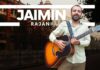 Jaimin Rajani holding a guitar, standing in front of an iconic Kolkata city backdrop, featuring historical architecture and a serene street view.