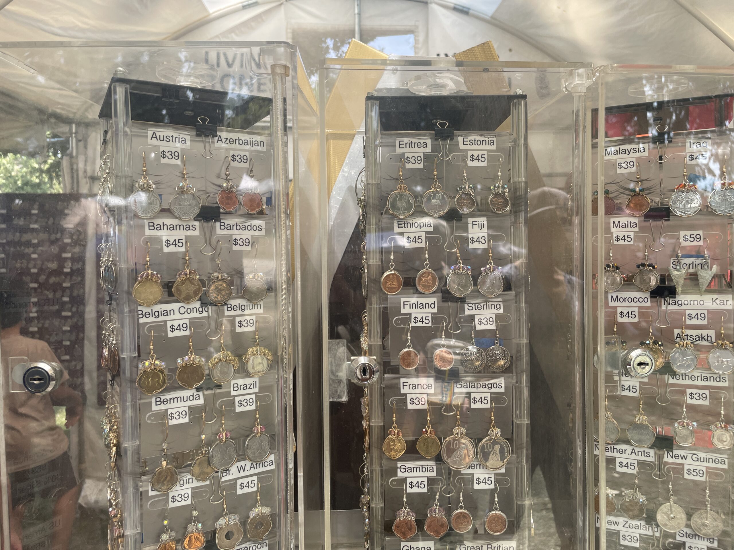 A collection of handmade coin pendants representing different countries, each meticulously crafted and displayed in a glass case. The pendants are labeled with prices and feature historical or cultural symbols from countries like Cuba, Ethiopia, and Bosnia.