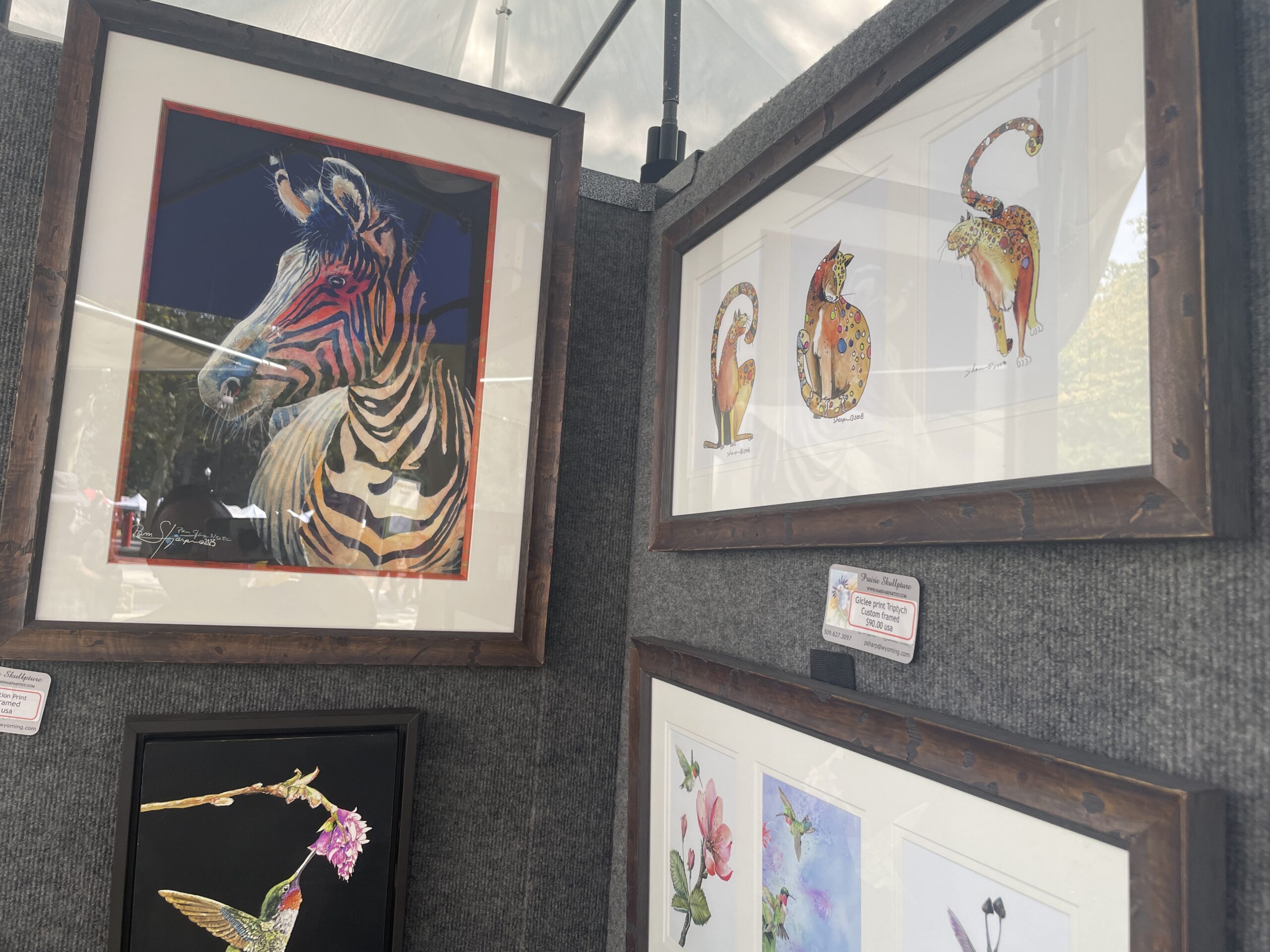 Framed artwork featuring a colorful zebra portrait and whimsical cat illustrations displayed at a booth in Art in the Park Boise 2024.