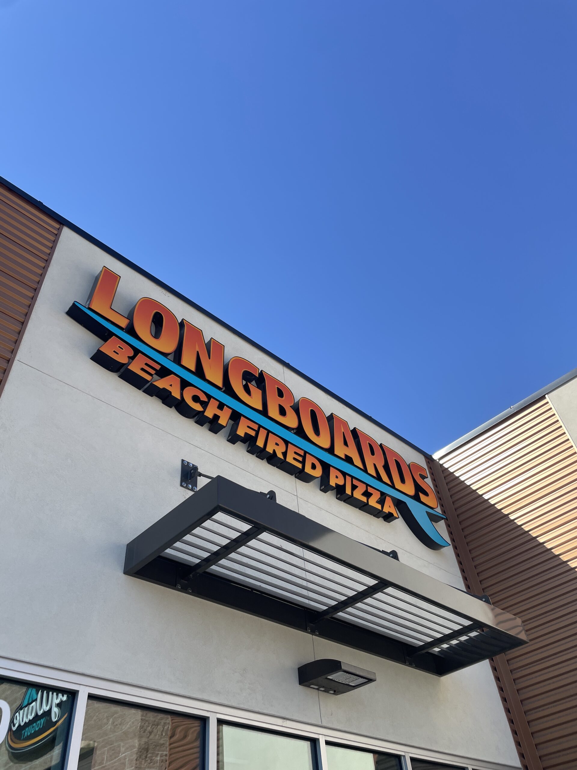 Exterior view of Longboards Beach Fired Pizza in Nampa, Idaho, featuring the vibrant orange and teal storefront sign against a clear blue sky.