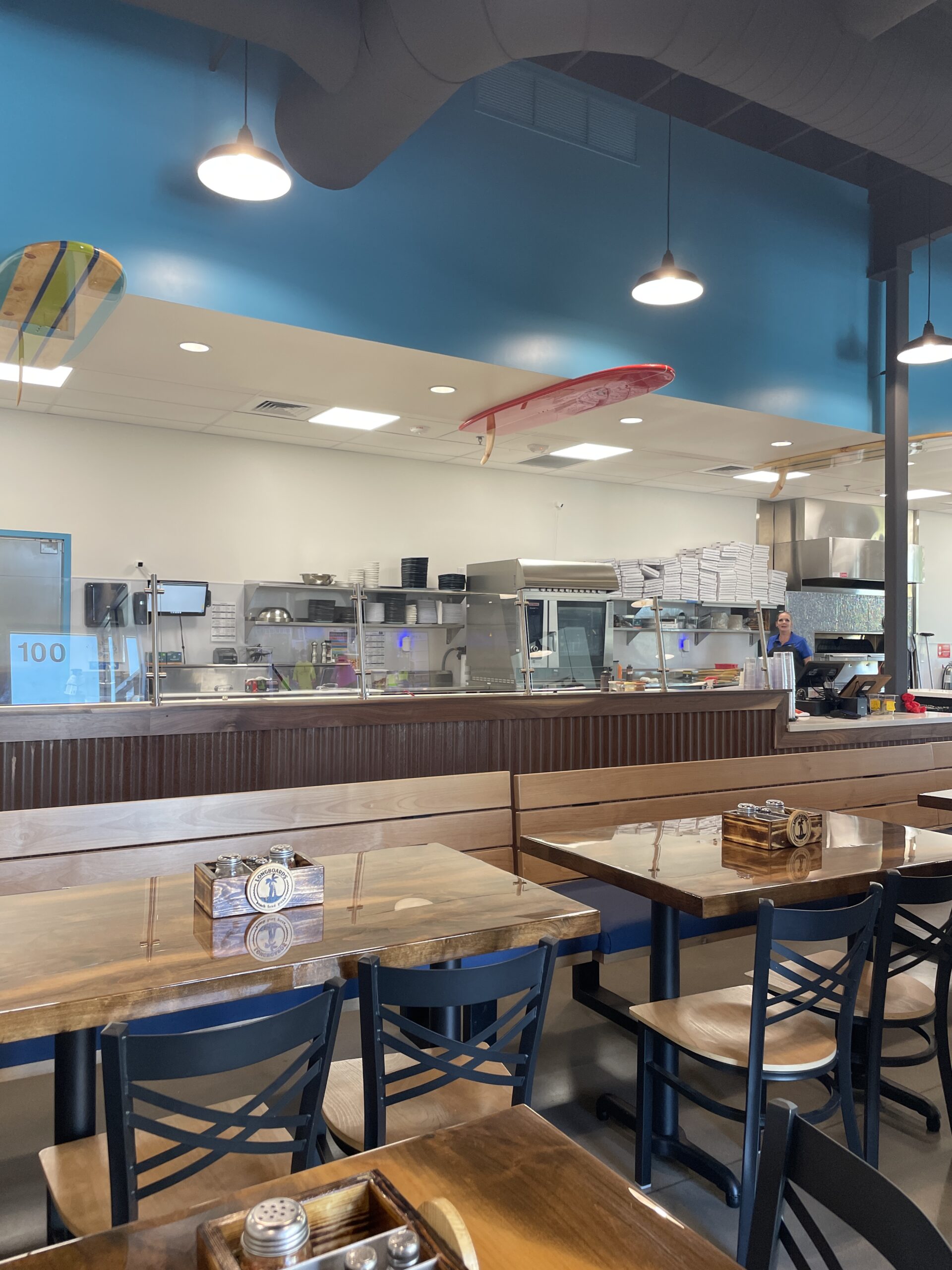 Dining area inside Longboards Pizza in Nampa, Idaho, featuring a view of the open kitchen, surfboard decor on the ceiling, and rustic wooden tables.