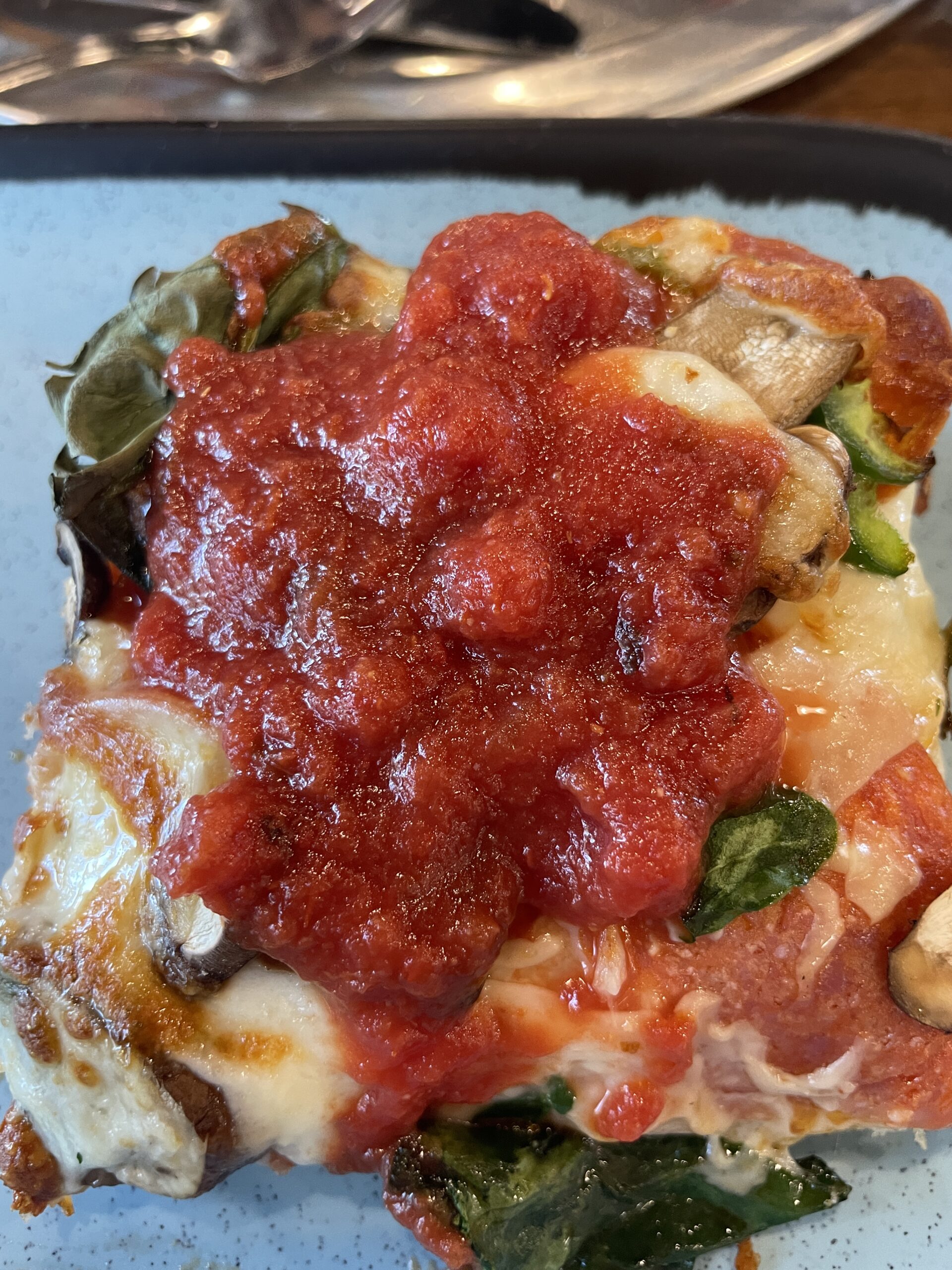 Close-up of a thick square slice of pizza topped with fresh spinach, mushrooms, and a generous layer of tomato sauce at Longboards Pizza in Nampa, Idaho.