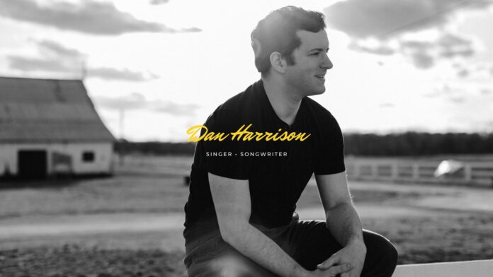 Dan Harrison, a Nashville-based singer-songwriter, sits outdoors in front of a barn wearing a black t-shirt. He is smiling and looking to his right, with a rural landscape in the background. The text 