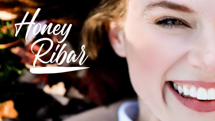 Close-up of Honey Ribar smiling with 'Honey Ribar' written in elegant script on the left side, featuring a blurred background with warm, earthy tones.