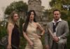 Rose Cora Perry and her band The Truth Untold posing in front of a stone castle. Rose stands center wearing an elegant beige dress, holding a guitar. To her left is the bassist, dressed in a dark dress with a visible tattoo on her shoulder, holding a bass guitar. On her right is the drummer, dressed in a gray suit, holding drumsticks with a relaxed stance. The band exudes a confident, edgy look against the backdrop of lush green trees and castle-like stone architecture.