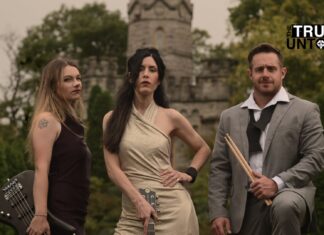 Rose Cora Perry and her band The Truth Untold posing in front of a stone castle. Rose stands center wearing an elegant beige dress, holding a guitar. To her left is the bassist, dressed in a dark dress with a visible tattoo on her shoulder, holding a bass guitar. On her right is the drummer, dressed in a gray suit, holding drumsticks with a relaxed stance. The band exudes a confident, edgy look against the backdrop of lush green trees and castle-like stone architecture.