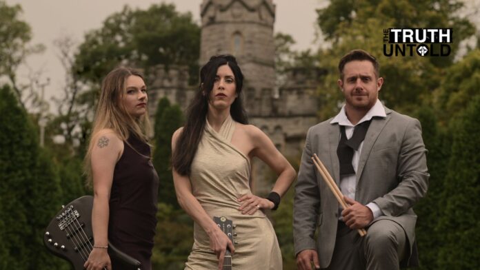 Rose Cora Perry and her band The Truth Untold posing in front of a stone castle. Rose stands center wearing an elegant beige dress, holding a guitar. To her left is the bassist, dressed in a dark dress with a visible tattoo on her shoulder, holding a bass guitar. On her right is the drummer, dressed in a gray suit, holding drumsticks with a relaxed stance. The band exudes a confident, edgy look against the backdrop of lush green trees and castle-like stone architecture.