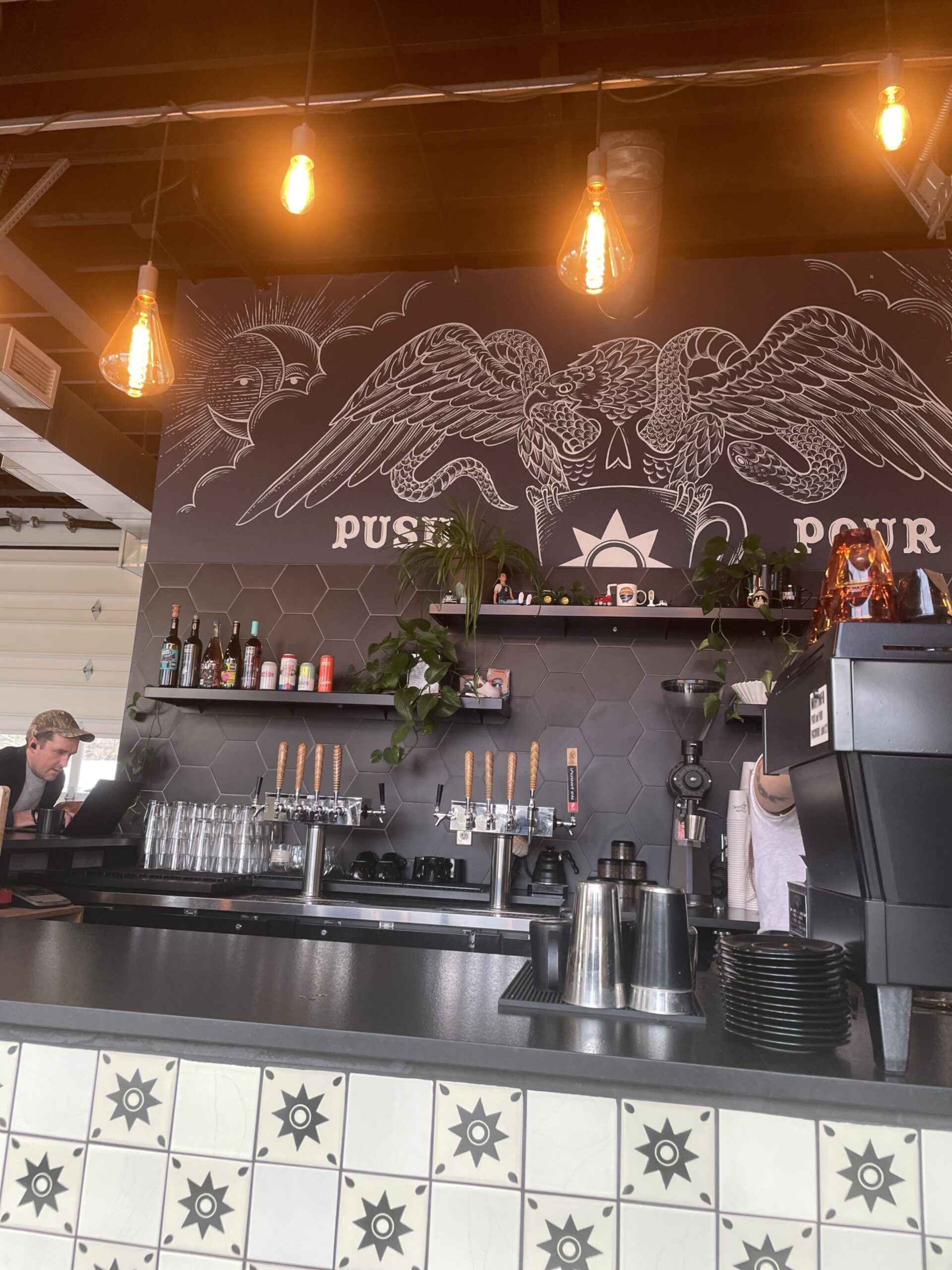 A modern coffee bar at Push and Pour featuring taps, coffee brewing equipment, and a large artistic mural with wings and a cup design.