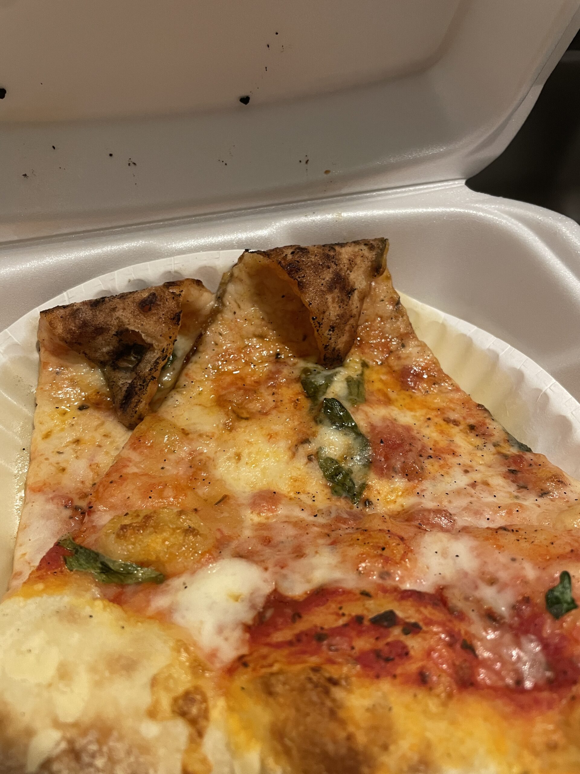 A slice of Guido’s basil pizza, with a thin, charred crust, melted mozzarella cheese, fresh basil leaves, and a vibrant tomato sauce, served in a takeout box.