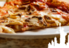 A close-up of a slice of New York-style pizza topped with fresh mushrooms, melted cheese, and tomato sauce, served on a white plate. The image features a stylized silhouette of the Statue of Liberty and New York City skyline in the bottom-right corner.