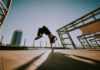 Breakdancer performing a handstand flip on an outdoor rooftop with a cityscape in the background.