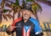 Marcus Hernandez of Orchestra Fuego smiling in front of a tropical cityscape, wearing a Puerto Rico-themed outfit and pointing to his band's logo.