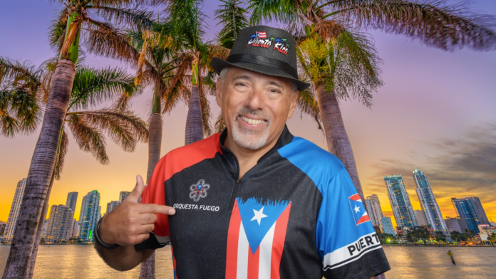 Marcus Hernandez of Orchestra Fuego smiling in front of a tropical cityscape, wearing a Puerto Rico-themed outfit and pointing to his band's logo.