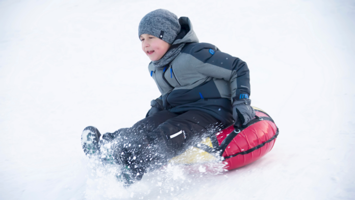 A young child in a gray winter jacket and beanie sledding down a snowy hill on a red inflatable tube, smiling with excitement.