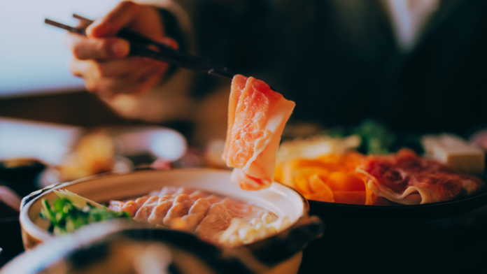 A person dipping thinly sliced beef into a steaming hot pot filled with fresh vegetables and broth at a Japanese shabu shabu restaurant.