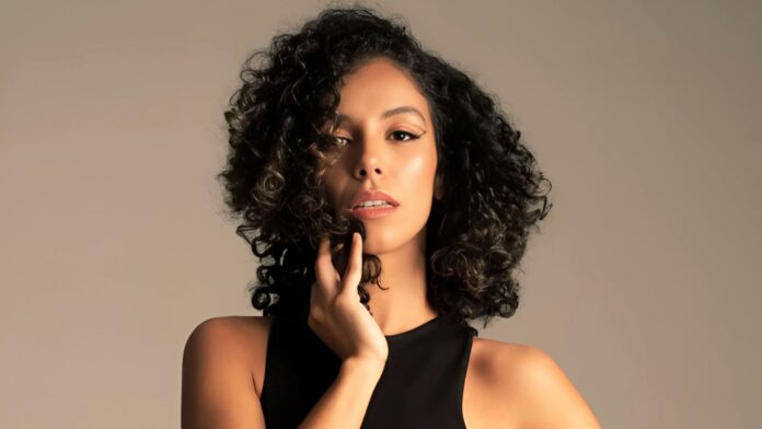 A portrait of Aria Fenix, a Colombian-born singer-songwriter, posing with her hand gently touching her face. She has voluminous curly hair, radiant skin, and wears a sleek black sleeveless top. Her expression is confident and captivating, with a soft, neutral background emphasizing her striking features.