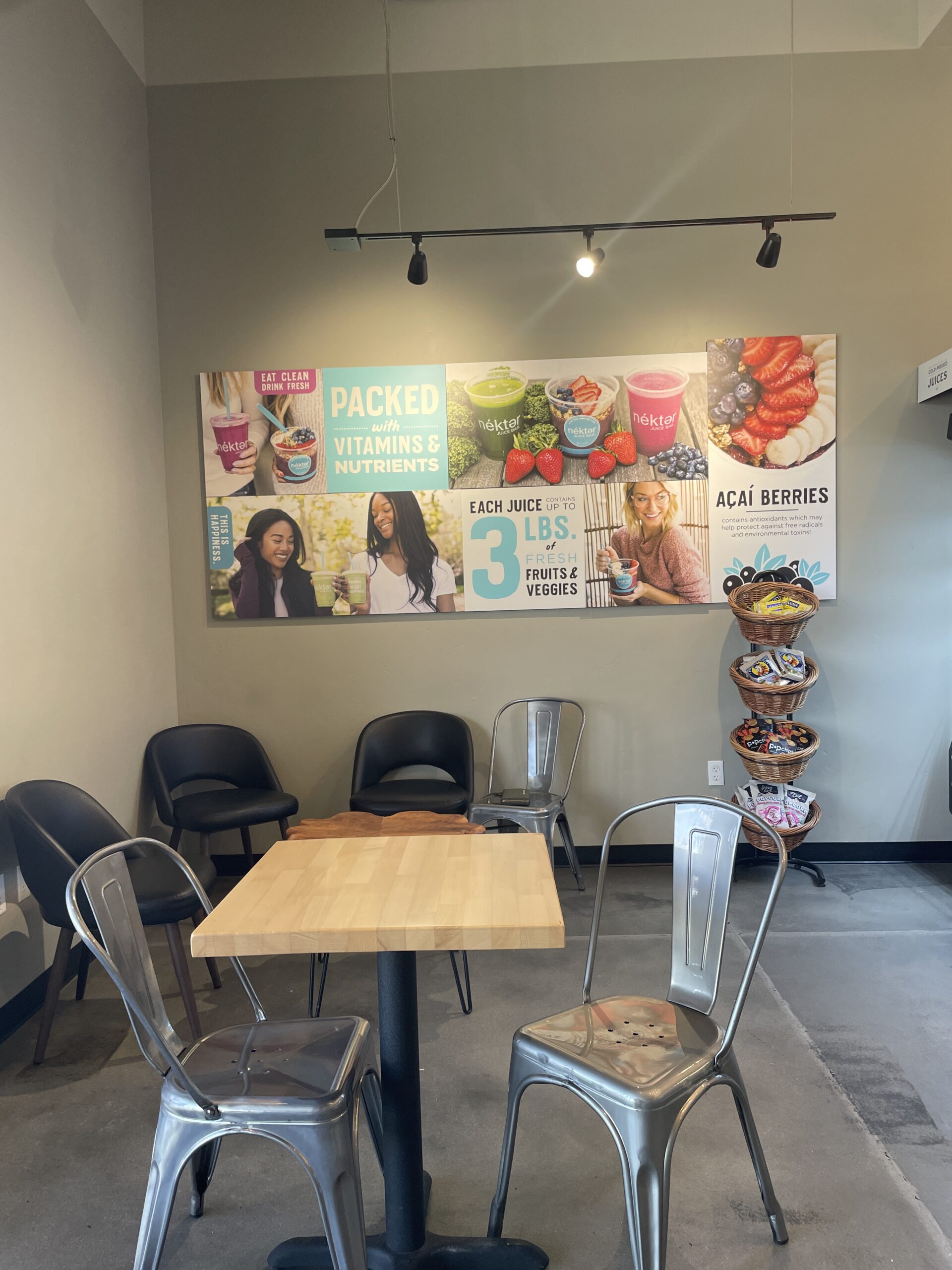 The cozy seating area inside Nekter Juice Bar in Meridian, Idaho, featuring black chairs, wooden tables, and a large mural promoting fresh juice and acai bowls.