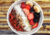 A vibrant acai bowl topped with goji berries, cacao nibs, shredded coconut, blackberries, bananas, and strawberries on a rustic wooden table.