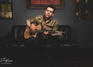 Bryan Copeland playing an acoustic guitar while seated on a black leather couch, eyes closed in deep musical expression.