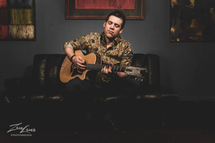 Bryan Copeland playing an acoustic guitar while seated on a black leather couch, eyes closed in deep musical expression.