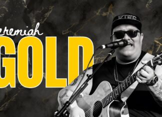 Jeremiah Gold performing live with guitar and microphone, wearing sunglasses and a hat that says “KING,” against a black marble background with bold yellow text.