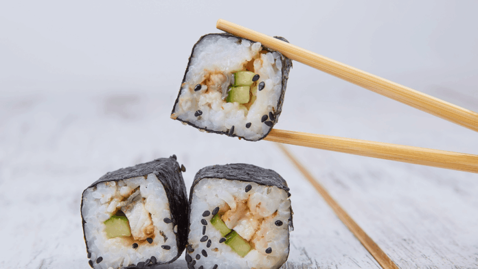 Sushi Bros Close-up of sushi rolls with cucumber, rice, and black sesame seeds held by chopsticks on a white wooden background.