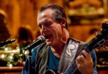 Jersey Johnny Flora: 45 Years of Guitar Mastery, Clapton Tribute & Original Blues Stories A passionate blues guitarist and singer performing live on stage with a microphone and electric guitar, set against warm ambient lighting and festive décor.