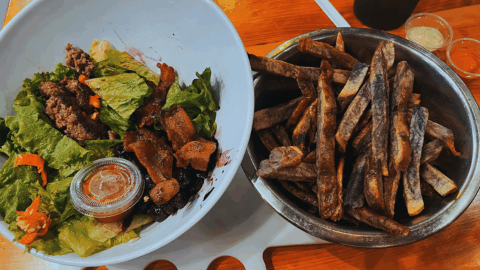 Boise Fry Company Fresh green salad topped with ground bison, crispy bacon, and blueberry compote, served alongside a metal bowl of thick-cut purple heirloom fries at Boise Fry Company.