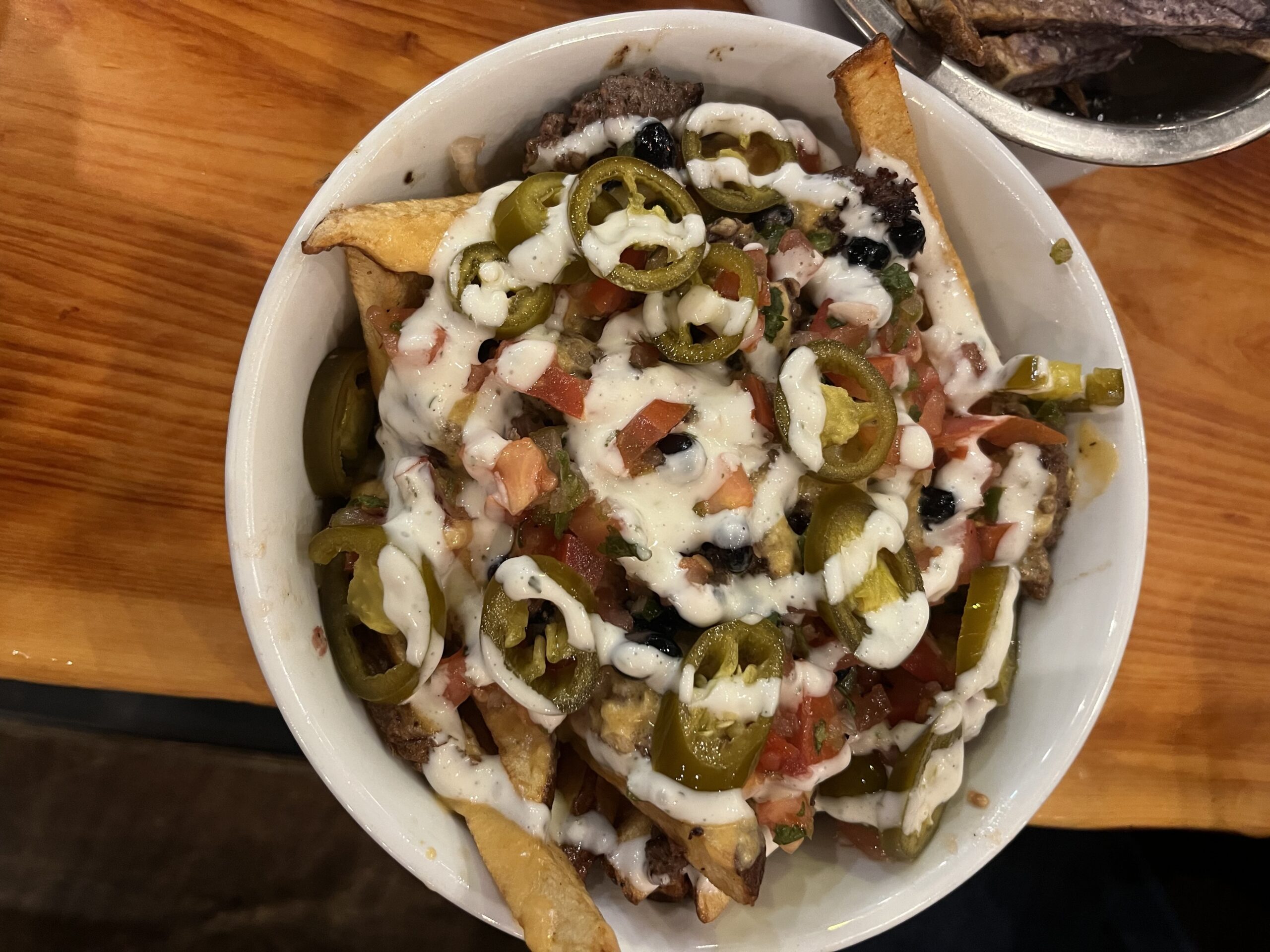 Loaded Boise Fry Company “Frychos” topped with jalapeños, diced tomatoes, black olives, and ranch drizzle over golden fries in a white bowl.