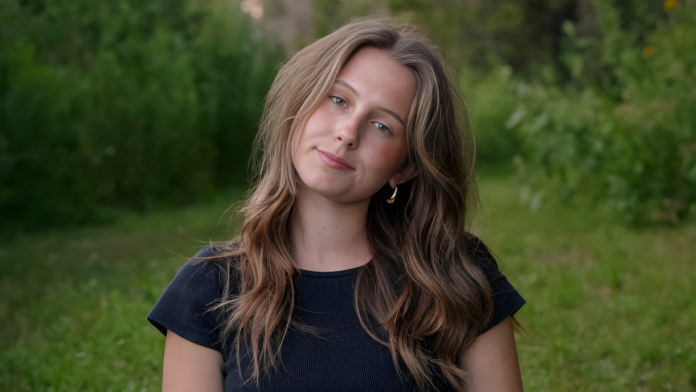 Close-up portrait of singer-songwriter Sydney Dale outdoors, with loose wavy hair, a black top, and a soft natural expression against a green, blurred forest background.