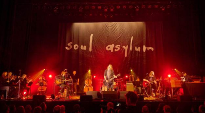 Dave Pirner on Soul Asylum’s Next Chapter & MPLS Unplugged Soul Asylum performs live onstage under red concert lighting, with guitars, strings and the band’s handwritten logo projected behind them.